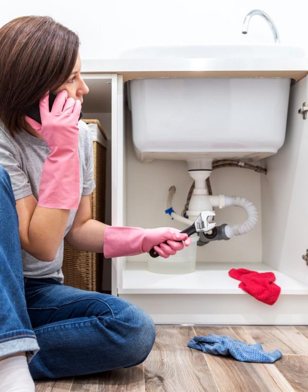 Woman wearing jeans, t-shirt, and pink rubber gloves holds a wrench in one hand and her phone in the other as she calls for help to fix her bathroom sink